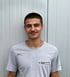 Ein junger Mann mit dunklem Haar, Schnurrbart und schwarzem T-Shirt lächelt.