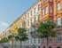 Reihe von bunten, historischen Wohngebäuden mit Bäumen und geparkten Autos an einer Straße unter blauem Himmel.