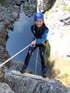 Lachende junge Frau in Neoprenanzug und Helm seilt sich über Felsen zu einem See ab.