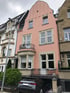 Rosa Gebäude mit geschwungenem Giebel, Erker, Balkon und einer Tiefgarageneinfahrt mit geparktem schwarzem Auto.