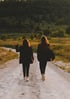 Zwei Frauen in dunkler Kleidung gehen auf einem Feldweg mit Bäumen und Gras im Hintergrund.