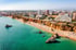 Aerial view of a crowded sandy beach with turquoise ocean, rock formations, and a city skyline.