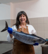 A smiling woman in a white shirt and brown apron holds a large tuna fish in blue-gloved hands.