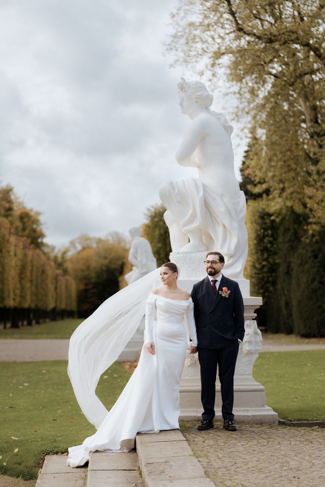 Brautpaar vor einer Skulptur im Schlosspark von Schloss Benrath in Düsseldorf, der Schleier weht im Wind.