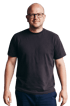 Ein Mann mit Glatze und Brille trägt ein schwarzes T-Shirt und eine blaue Hose, lächelt leicht.