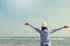 Frau mit Hut und ausgebreiteten Armen steht am Meer unter blauem Himmel.