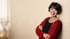 Hintergrundbild. Andrea Martha Becker  (offene rote Hoodiejacke, schwarzes Topp), rechts stehend mit verschränkten Armen, vor cremefarbener Wand, links ein leicht wehender lichtdurchlässiger heller Vorhang, davor eine cremefarbene Vase mit Trockenblumen. Besonderes Merkmal: zugewandtes Lachen