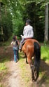 Eine Person reitet auf einem braunen Pferd durch einen Wald, während eine zweite Person daneben herläuft.