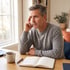 Middle-aged man in a grey sweater sits at a desk with an open notebook, pen, and mug, looking thoughtfully out a window.