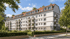 Modernes Mehrfamilienhaus mit weißer Fassade, Balkonen und rotem Satteldach vor blauem Himmel mit Bäumen im Vordergrund.