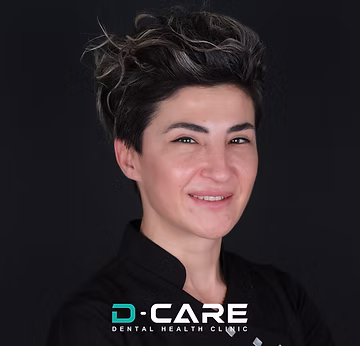 Eine lächelnde Frau mit kurzen, dunklen Haaren und hellen Strähnen trägt ein schwarzes Oberteil, darunter das Logo "D-CARE DENTAL HEALTH CLINIC".