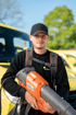 Ein Gärtner mit schwarzer Kappe und Rucksacktragesystem hält einen orangefarbenen Husqvarna Laubbläser vor einem gelben Auto.