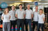 Sechs Fitnesstrainer in hellgrauen Poloshirts und Sportbekleidung stehen lächelnd nebeneinander in einem Fitnessstudio.