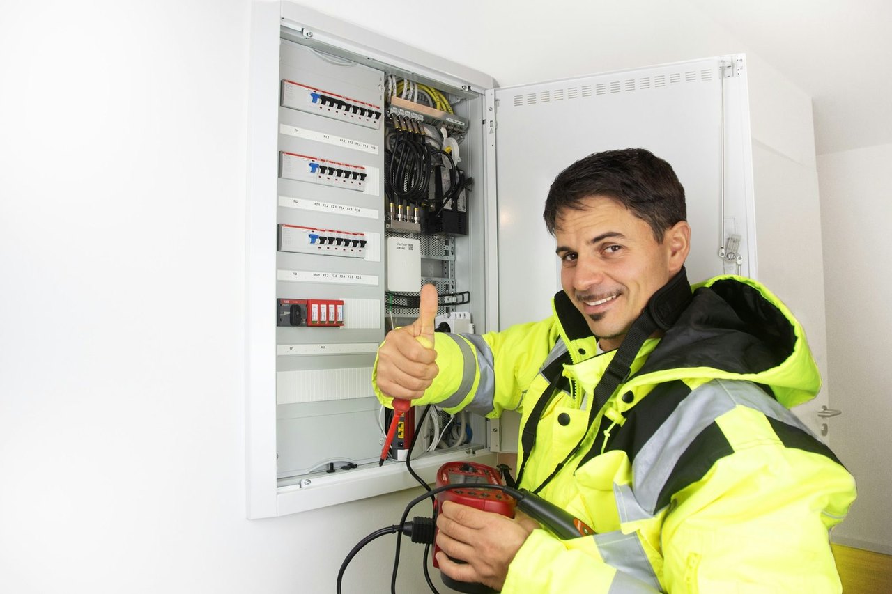 Elektriker in Warnjacke vor einem geöffneten Zählerschrank mit Messgerät in der Hand