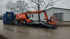 Ein blauer LKW transportiert eine orangefarbene Gelenk-Arbeitsbühne auf einem Tieflader.