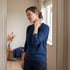 A woman in a blue sweater stands indoors, grimacing while holding her hand to the side of her neck.