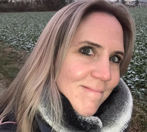 Frau mit blondem Haar und gestreiftem Schal lächelt vor einem Feld mit leichtem Schnee.