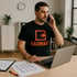 Mann im schwarzen T-Shirt mit "RAUMAKT"-Logo telefoniert am Laptop mit Raumplänen  auf dem Tisch.
