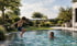 Ein Junge springt in einen Gartenpool, während eine Frau im Wasser wartet; im Hintergrund eine grüne Gartenanlage.