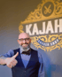 Ein lächelnder Mann mit Bart, Brille, Weste und Fliege zeigt den Daumen hoch vor dem Logo „KAJAH BARBER SHOP“.