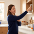 A woman in a navy sweater winces while holding her shoulder and reaching for a glass jar in a kitchen cabinet.