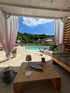 Überdachte Terrasse mit Holzmöbeln und Blick auf einen Gartenpool unter blauem Himmel.