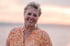 Eine lächelnde Frau mit kurzem grauem Haar und orangefarbenem Hemd vor einem verschwommenen Sonnenuntergang am Meer.