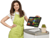 Lächelnde Mitarbeiterin in grüner Schürze steht neben einem POS-Kassensystem mit Touchscreen-Monitor.