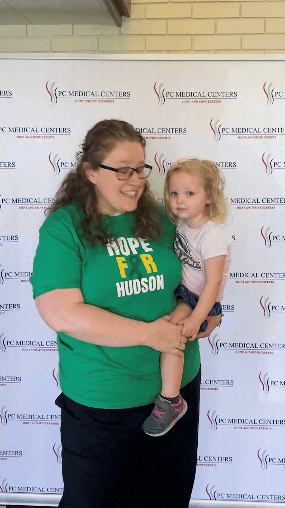 Woman in a green "Hope for Hudson" shirt holding a toddler in front of the PC Medical Centers backdrop