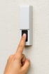Ein Zeigefinger drückt einen schwarzen Knopf an einem weißen Türlautsprecher auf einer strukturierten Wand.