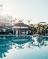 A resort hotel with white buildings, palm trees, and a swimming pool reflecting the sky and surroundings.