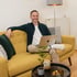 Ein lächelnder Mann sitzt mit einem MacBook auf einem gelben Sofa im Homeoffice.