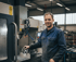 Ein Mann in blauer Arbeitskleidung und Schutzbrille lächelt neben einer CNC-Maschine.