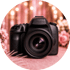Schwarze DSLR-Kamera mit großem Objektiv, umgeben von rosa Blumen und Bokeh-Lichtern.