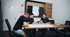 Drei Männer in schwarzer Kleidung sitzen an einem Holztisch und besprechen Baupläne in einem modernen Büro.