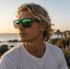 Blonder Mann trägt eine grüne Sonnenbrille vor einem sonnigen Strand bei Sonnenuntergang.