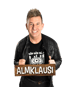 Almklausi lächelt in schwarzer Lederjacke und hält ein Holzschild mit seinem Namen vor schwarzem Hintergrund.