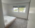 Leeres Schlafzimmer mit grauem Teppich, weiß gestrichenen Wänden, einem Fenster und einem Bett mit Plastikbezug.