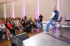 Speaker in denim jacket on a stool addressing an audience seated in rows, pink uplighting on walls.