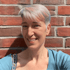 Eine Frau mit kurzen, grau-blonden Haaren und einem blauen Oberteil lächelt vor einer roten Ziegelwand.