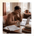 A woman sits at a desk with a laptop, books, and coffee, resting her head on her hand with a tired expression.