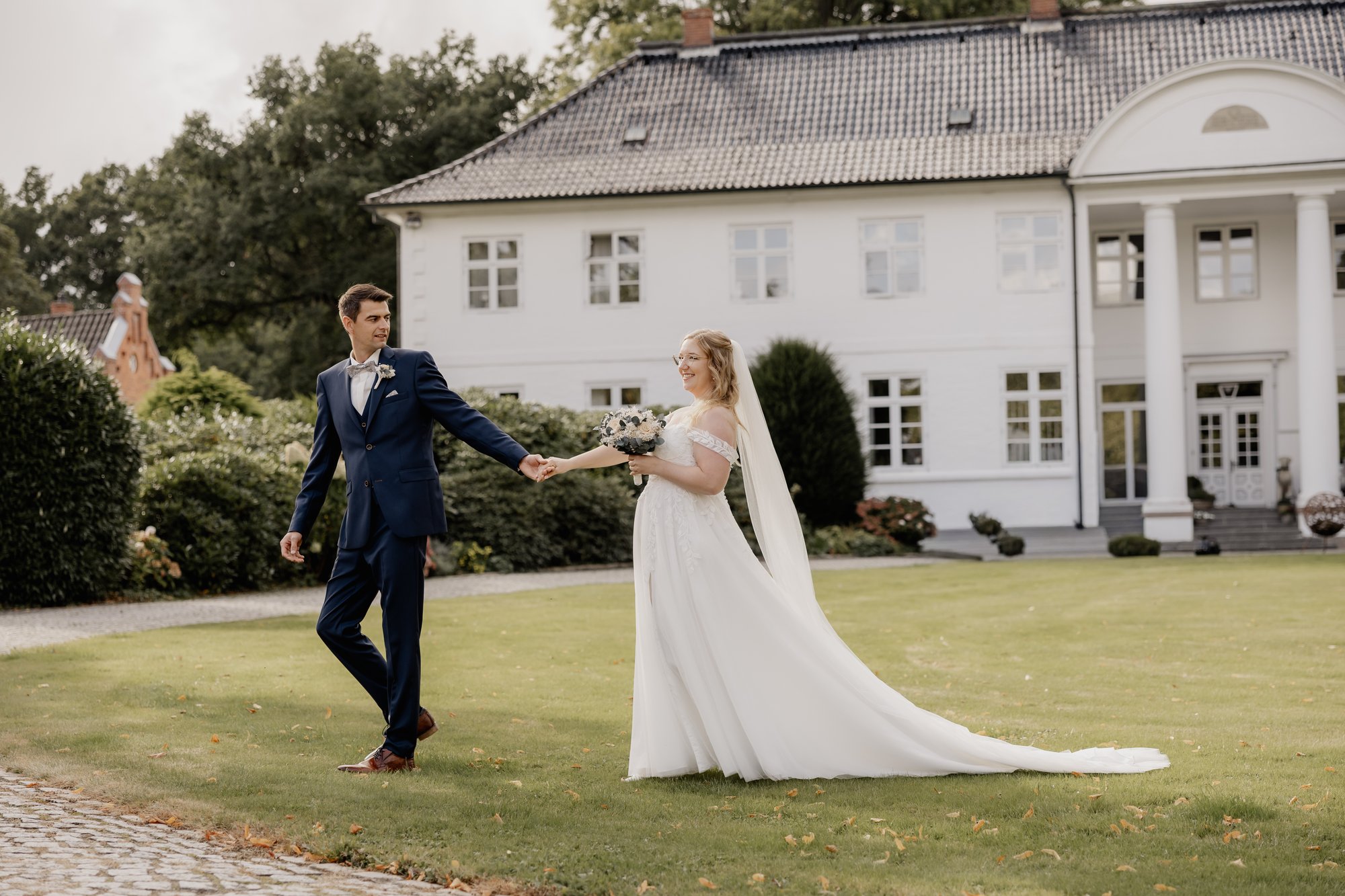 Brautpaar geht Hand in Hand über den Rasen vor einem weißen Gutshaus