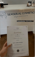 Hand hält ein Zertifikat für "Lash- und Browstylistin" von Semseral Academy vor einem Schreibtisch mit "SEMSERAL COSMETICS style and more" Schriftzug.