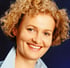 Eine lächelnde Frau mit blonden Locken und einem hellblauen Hemd vor dunklem Hintergrund.
