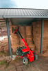 Roter Manitou-Teleskoplader mit ausgefahrenem Arm stapelt Heuballen unter einem Metallschuppen.