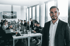 Mann lächelt im Vordergrund, dahinter ein Büro mit Mitarbeitern, die an Computern arbeiten.