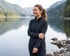 A woman in a charcoal fleece jacket stands by a calm lake with forested mountains in the background.