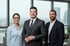 Drei lächelnde Geschäftsleute, zwei Männer und eine Frau, stehen in einem modernen Büro vor einem Fenster.