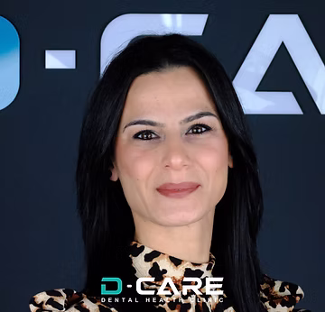 Eine junge Frau mit langen dunklen Haaren und einem Leopardenmuster-Oberteil lächelt vor einem „D-CARE DENTAL HEALTH CLINIC“-Logo.