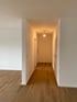 Flur mit Holzboden, weißen Wänden und einer Deckenlampe am Ende des Gangs.